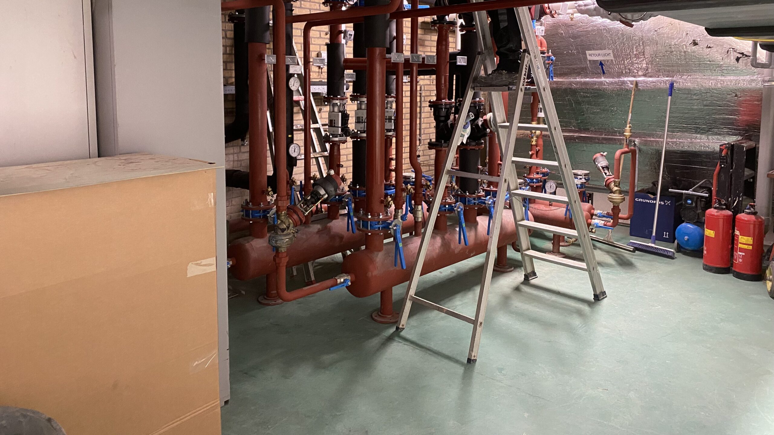 A view of a mechanical room featuring various pipes and valves in red and blue, along with a ladder and storage box in the foreground.